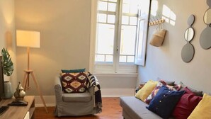 A living room with a grey sofa, a wooden floor, a standing lamp, a chair with patterned cushions, and a window with blinds.