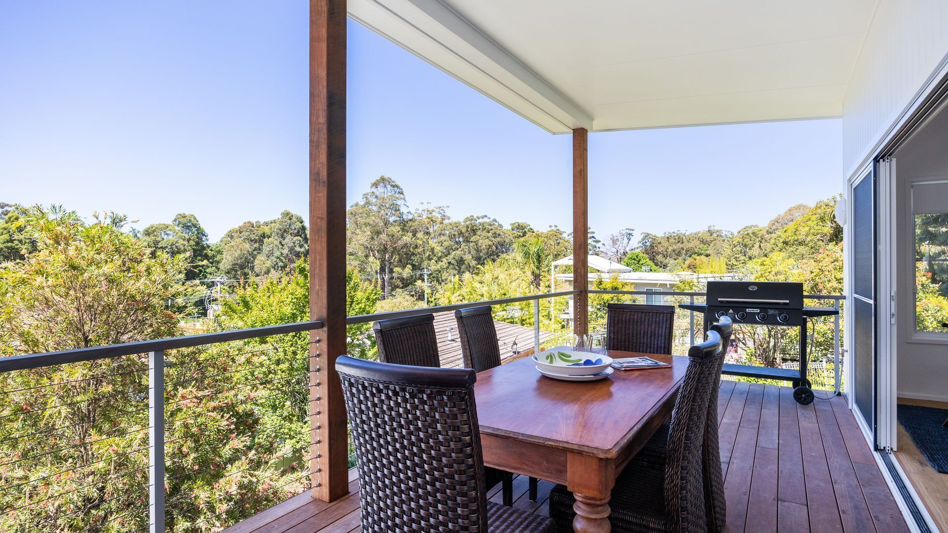 Un balcon avec une table et des chaises en bois, un barbecue et une vue sur une zone boisée.