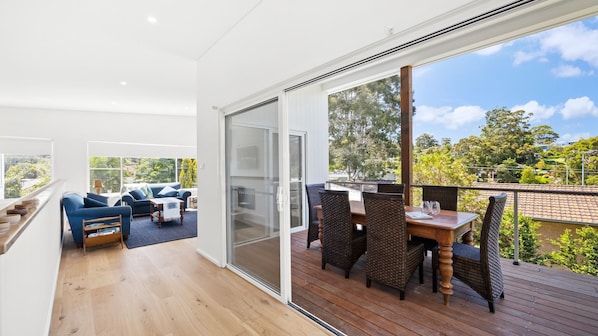 A spacious balcony with a wooden deck, a dining area with wicker chairs, and a sliding glass door leading to a living area with a blue sofa and a coffee table. - Bombora Pearl in Mollymook (Mollymook Beach)