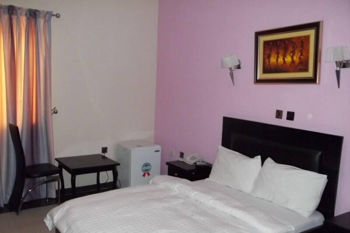 A hotel room with a bed, a desk, a chair, a small table, a nightstand, a lamp, and a painting on the wall.