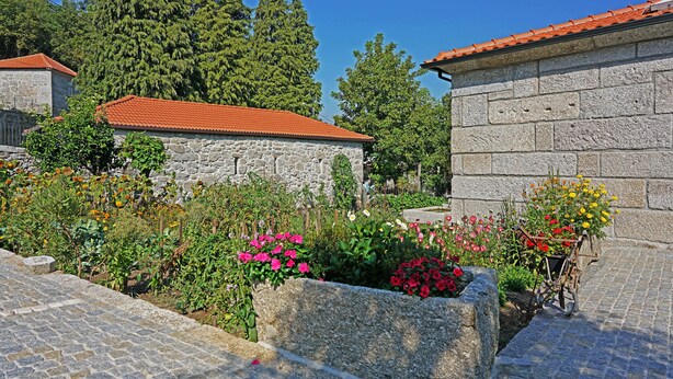 A stone building with a red-tiled roof, surrounded by a garden with various flowers and a wheelbarrow. - Porch House (Braga)