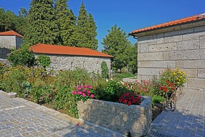 A stone building with a red-tiled roof, surrounded by a garden with various flowers and a wheelbarrow.
