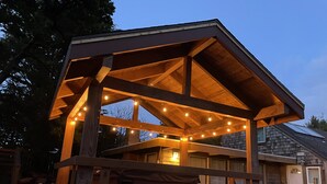 A wooden gazebo with string lights, a bench, and a view of a house in the background. - Beach Retreat House w/Ocean Views, Covered Hot Tub, & Large Fenced Yard (Lincoln City)