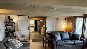 A living room with a grey sofa, a black leather sofa, and a wooden coffee table. There is a kitchen in the background with a fridge and cabinets. - Beach Retreat House w/Ocean Views, Covered Hot Tub, & Large Fenced Yard (Lincoln City)