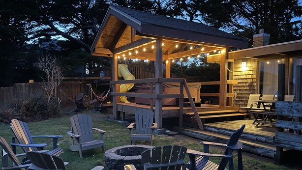 A wooden deck with Adirondack chairs, a fire pit, and a covered seating area with string lights. - Beach Retreat House w/Ocean Views, Covered Hot Tub, & Large Fenced Yard (Lincoln City)