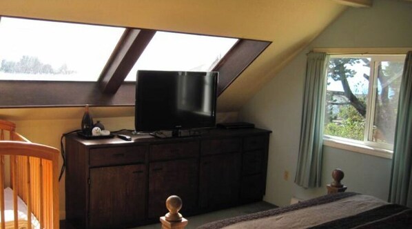 A room with a sloped ceiling, a television on a wooden cabinet, a window with curtains, and a small wooden chair. - Beach Retreat House w/Ocean Views, Covered Hot Tub, & Large Fenced Yard (Lincoln City)