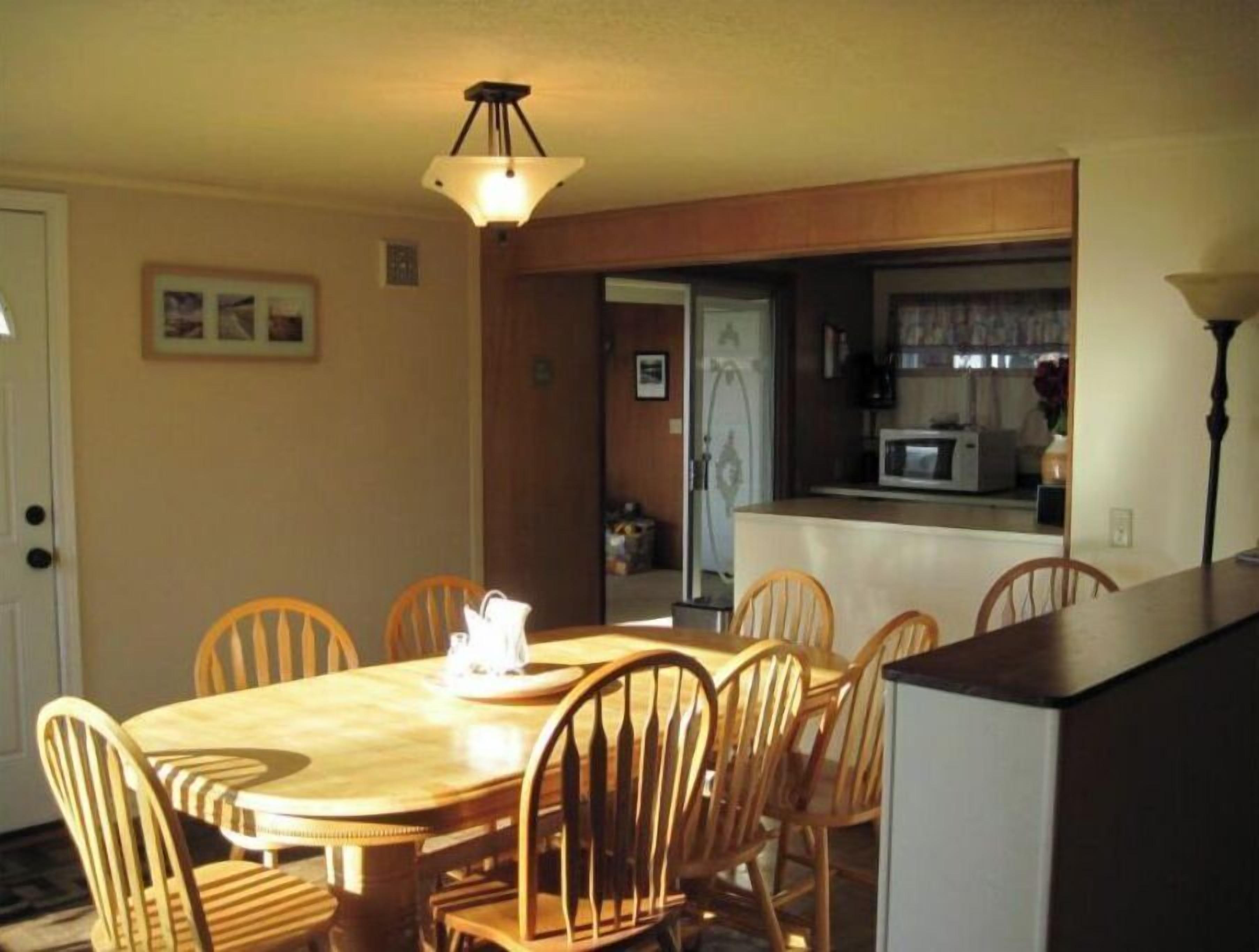 A dining area with a wooden table and chairs, a hanging light fixture, and a view into the kitchen.