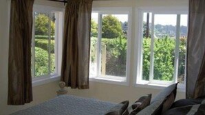A bedroom with a bed, curtains, and a view of greenery outside. - Beach Retreat House w/Ocean Views, Covered Hot Tub, & Large Fenced Yard (Lincoln City)