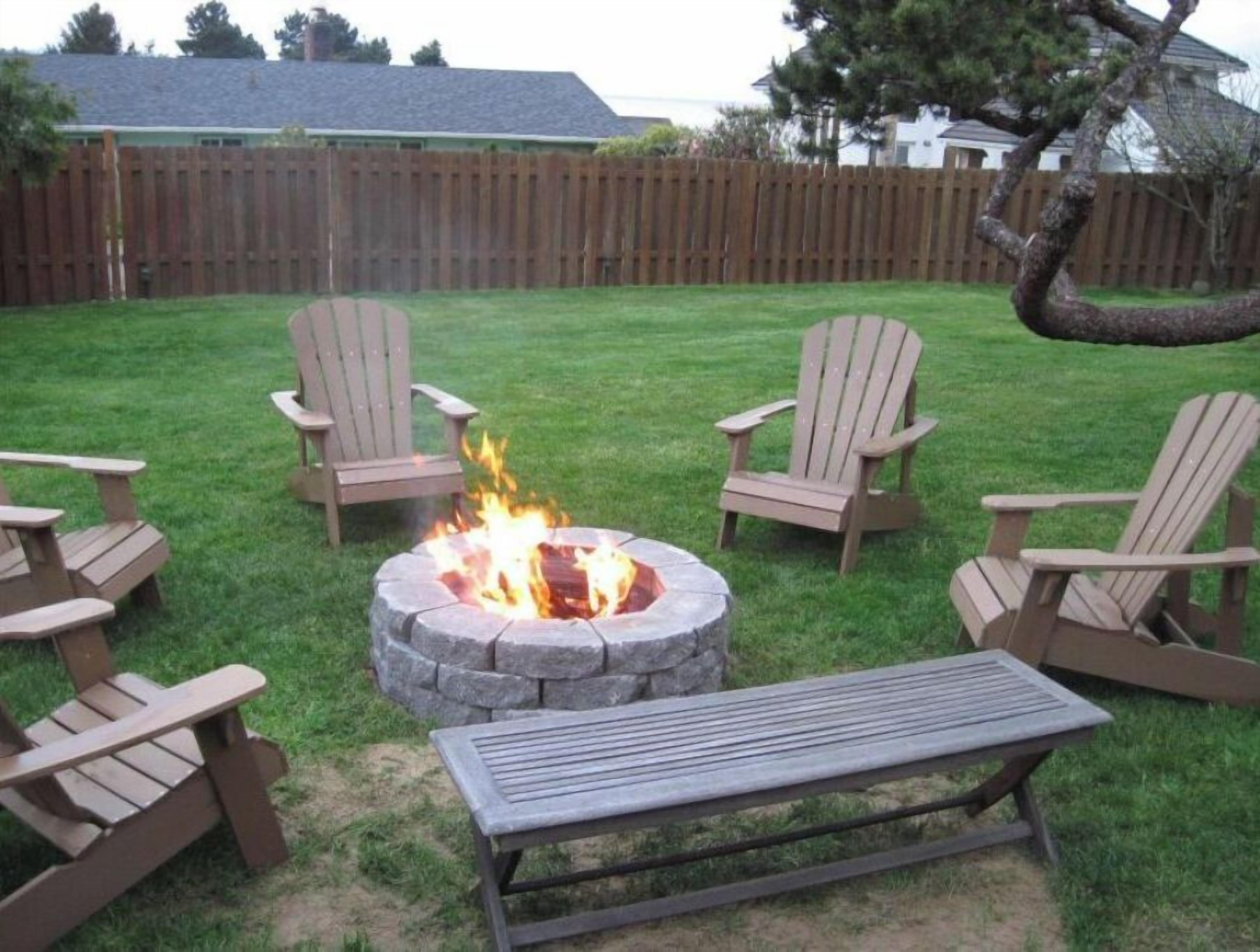 Een achtertuin met een vuurkorf van steen, omringd door houten Adirondack-stoelen en een bank.