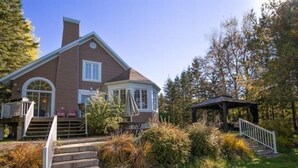 A large house with a covered porch, a staircase leading to the entrance, and a small gazebo in the yard. - Waterfront cottage Lac Quenouille (Val-des-lacs)