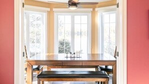 A dining area with a wooden table and bench, large windows, and a ceiling fan. - Waterfront cottage Lac Quenouille (Val-des-lacs)