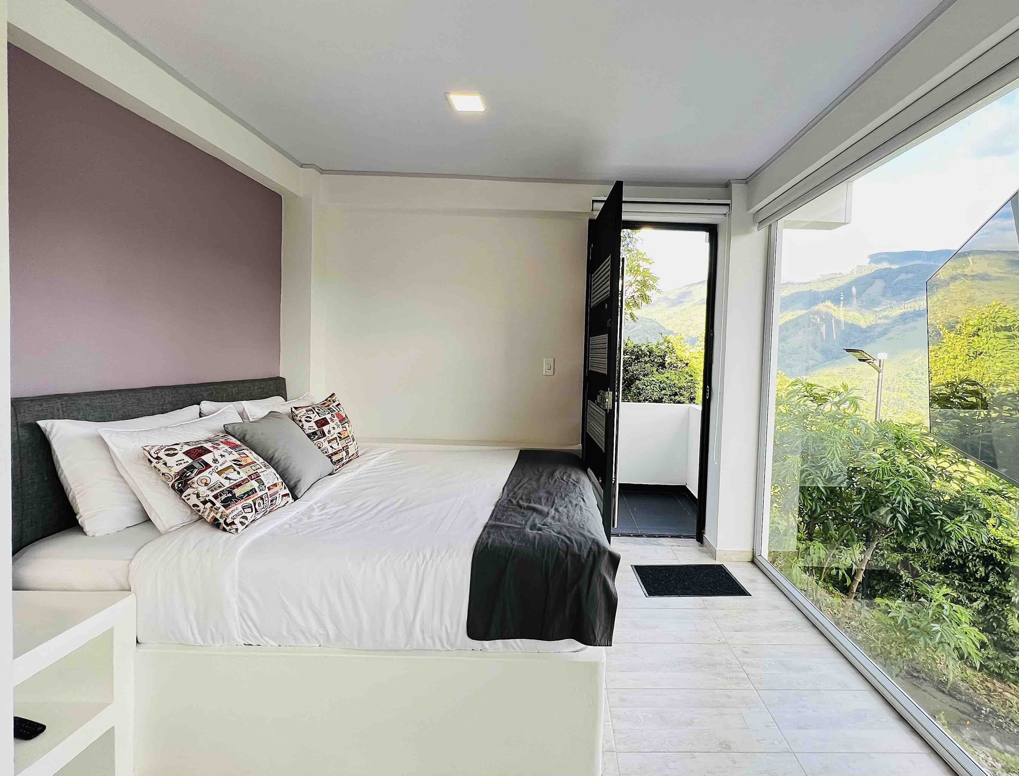 Een moderne slaapkamer met een groot bed, uitzicht op groen en bergen door een glazen deur, en een klein balkon.