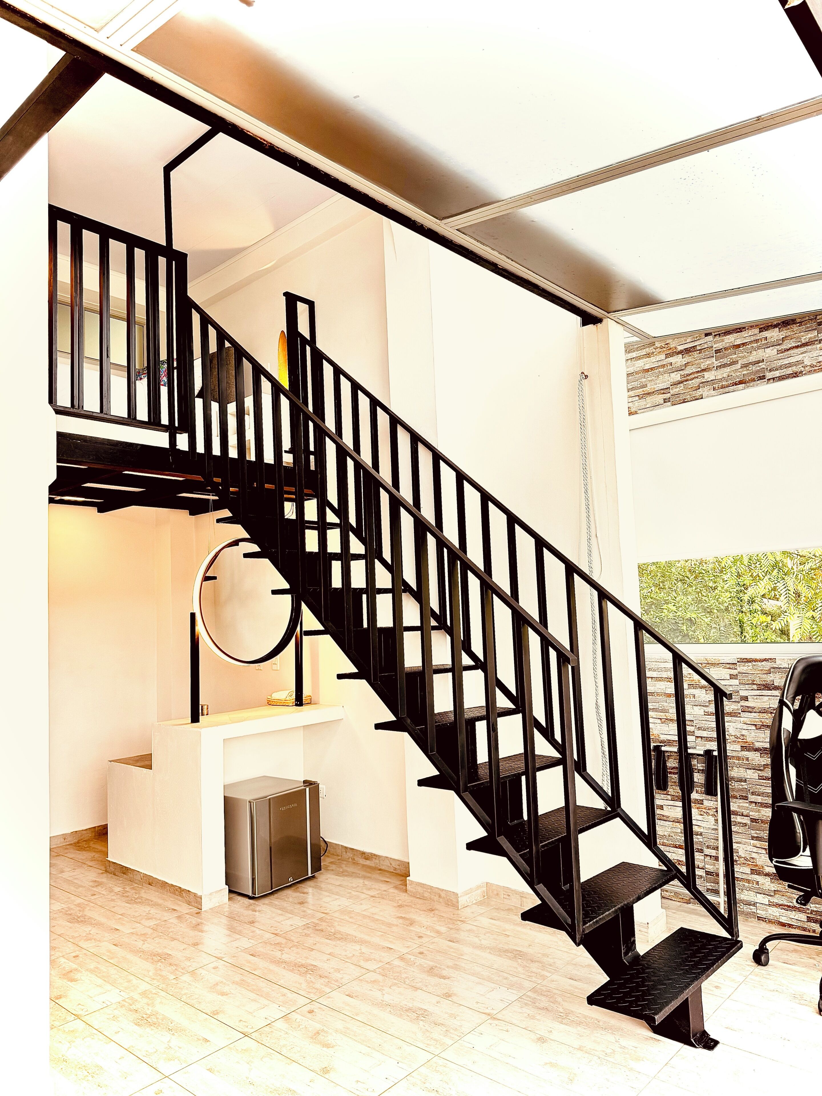 Un interior moderno con una escalera de metal negro, una pequeña zona de cocina y vistas a la vegetación exterior.