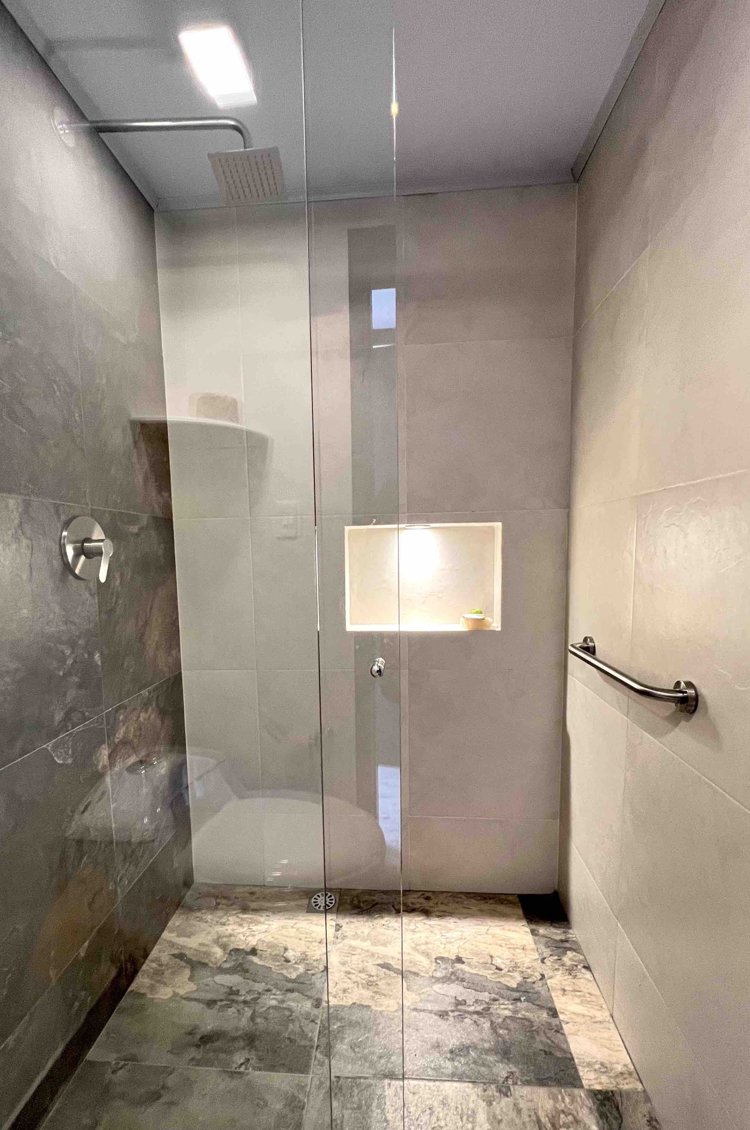 Un área de ducha moderna con puerta de cristal, una barandilla y un estante pequeño con luz.