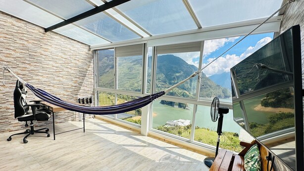 Una habitación con hamaca, escritorio y vistas a un río y montañas a través de amplios ventanales.