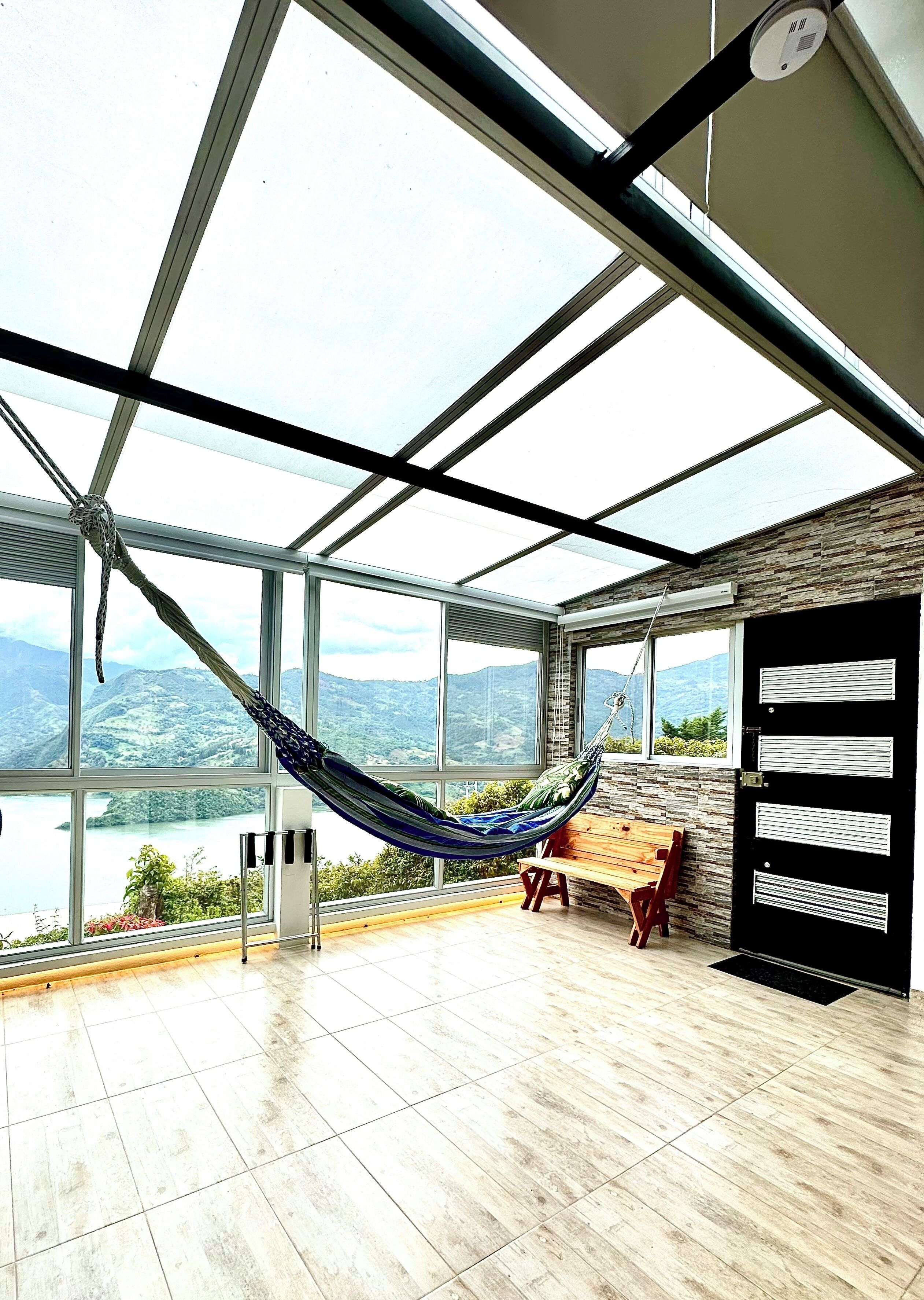 A spacious room with large windows offering a view of a lake and mountains. A hammock is suspended from the ceiling, and there is a wooden bench near the windows.