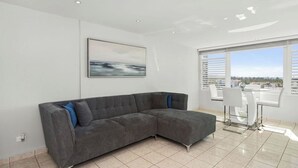 Un salon moderne avec un canapé d’angle gris, un coin repas avec des chaises blanches et une grande fenêtre donnant sur la ville.
