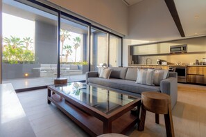 Een moderne woonkamer met een glazen salontafel, een grijze bank met witte kussens en een houten kruk.