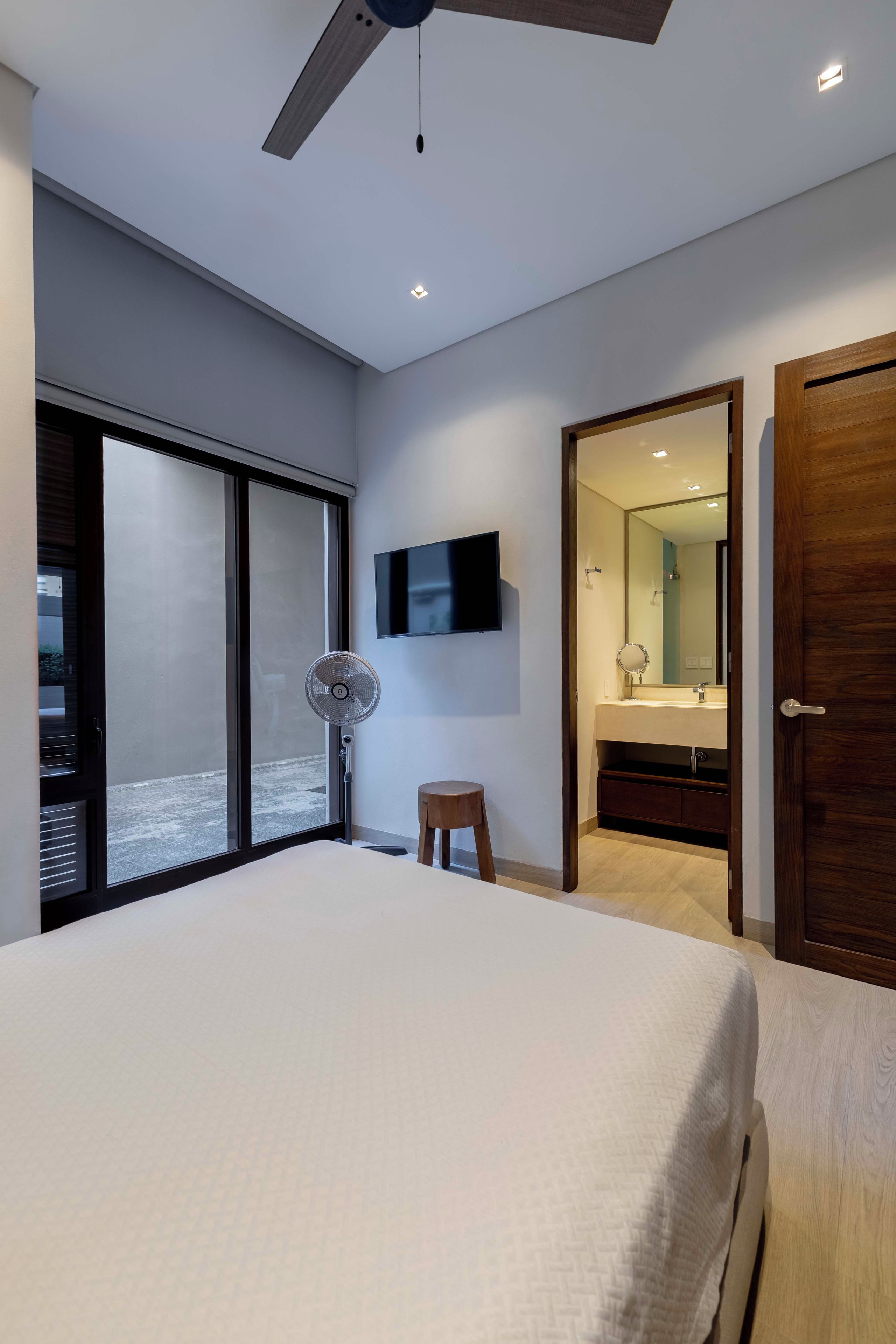 Ein modernes Schlafzimmer mit einem großen Bett, einem Deckenventilator, einem Fernseher und einer Glastür, die auf einen Balkon führt.