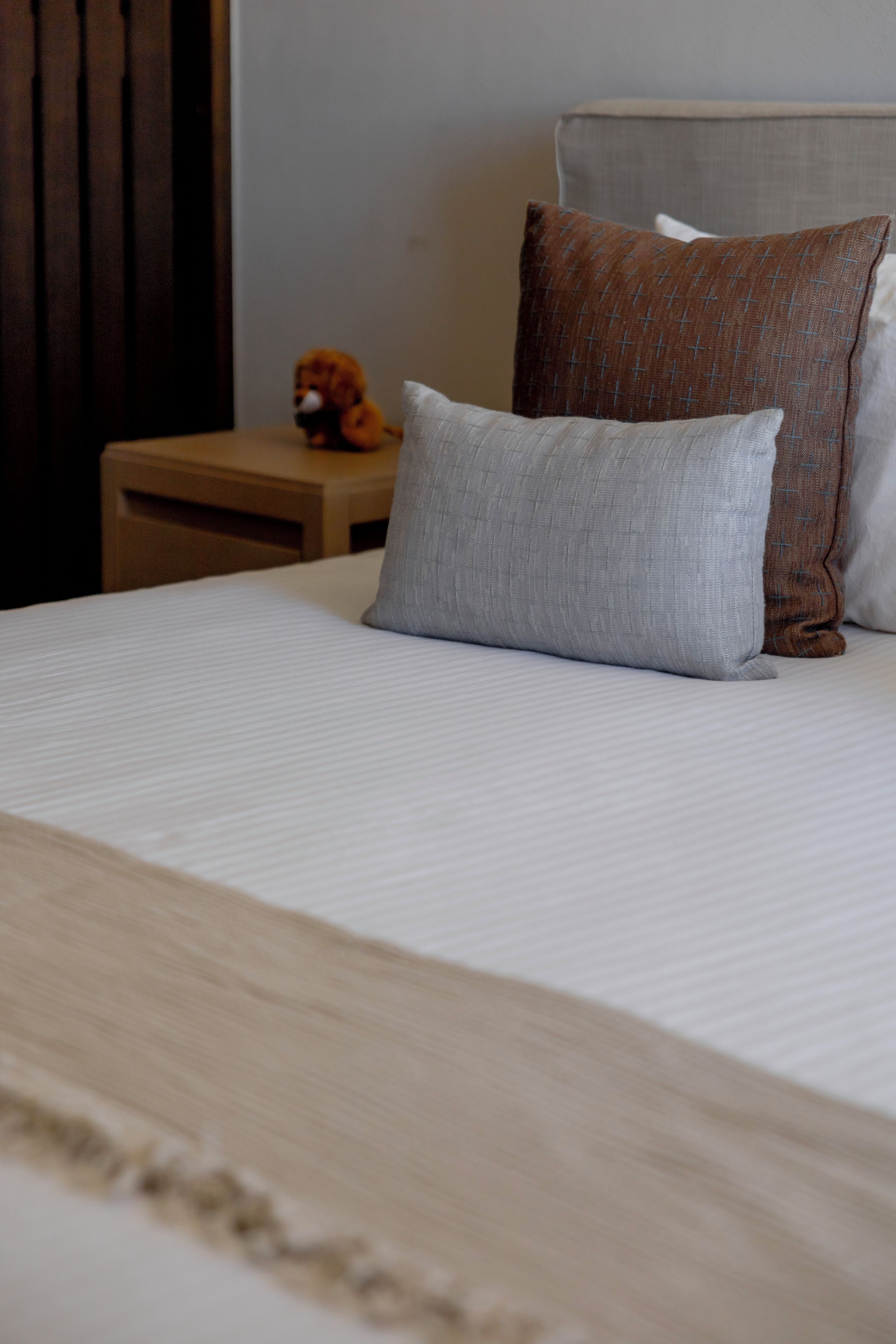 Een keurig opgemaakt bed met een wit laken en twee kussens, één grijs en één bruin met een kruispatroon.