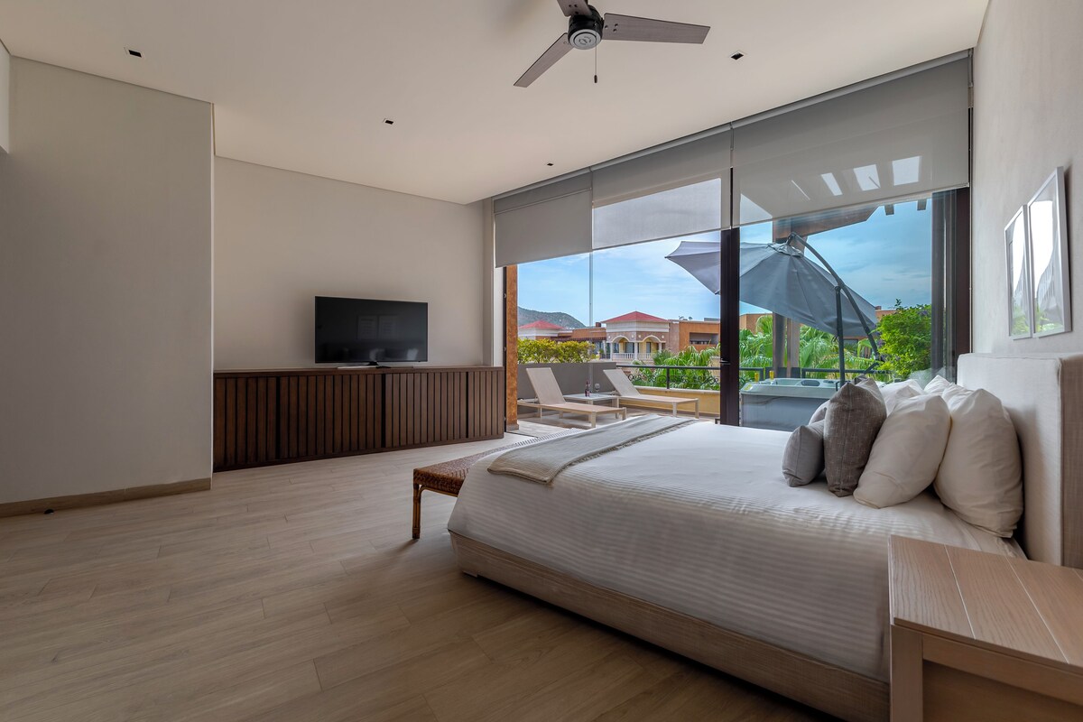 Une chambre moderne avec un grand lit, une télévision et un balcon offrant une vue sur les bâtiments et la verdure.