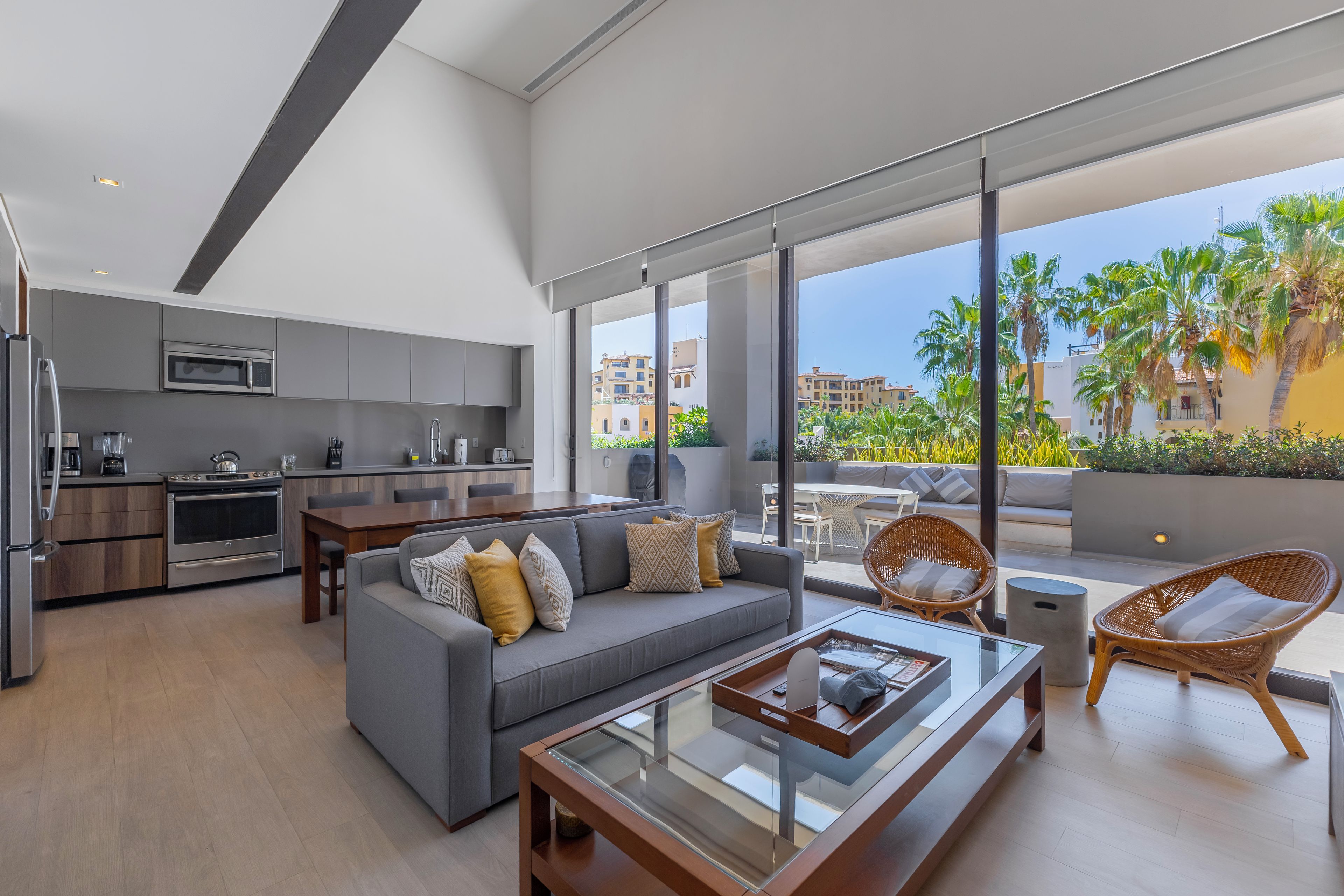 A modern living space with a kitchen, a gray sofa with patterned cushions, a glass-top coffee table, and wicker chairs.