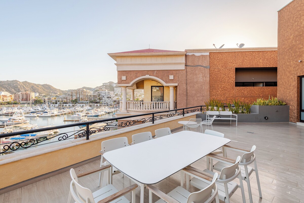 Um terraço com mesa e cadeiras brancas, dando vista para uma marina com barcos.