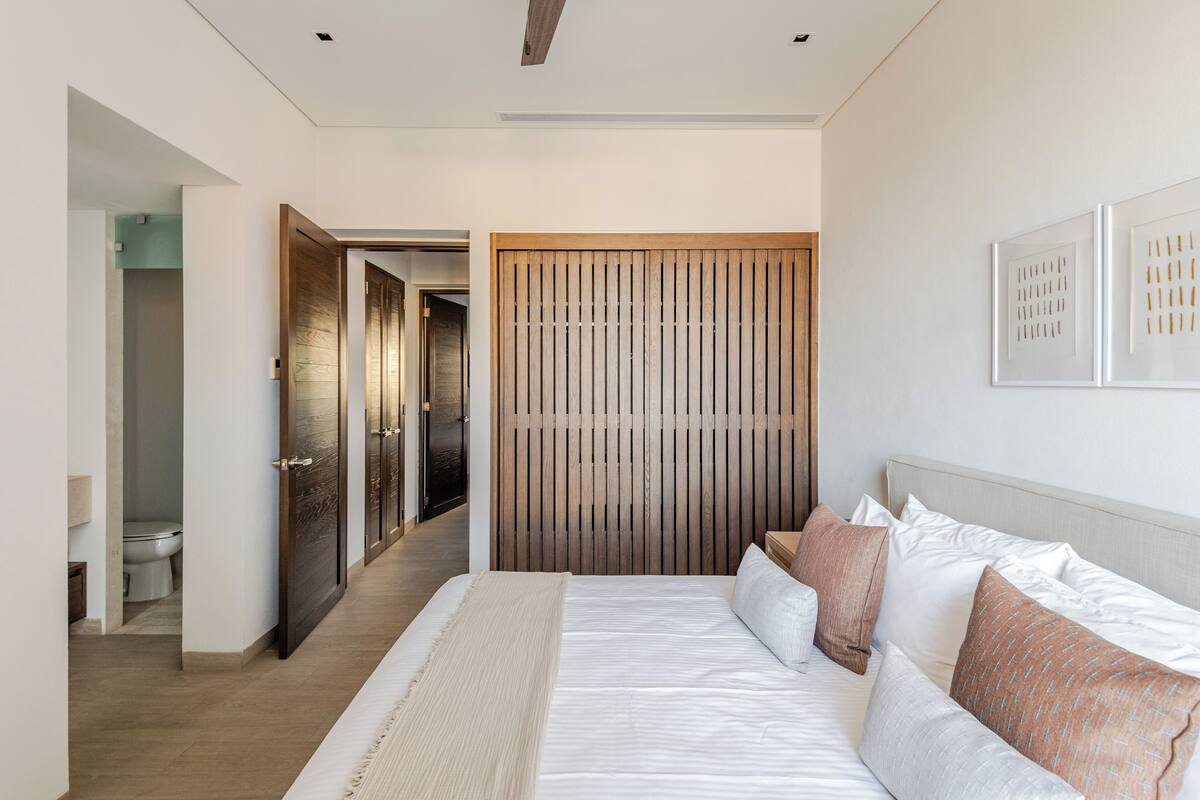 Une chambre moderne avec un grand lit, une cloison en lattes de bois et un couloir menant à une salle de bain.