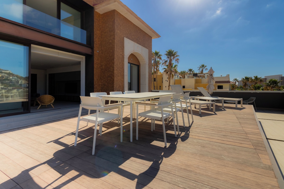Um pátio externo com deck de madeira, mesas e cadeiras brancas, e uma porta de vidro que dá para um edifício moderno.