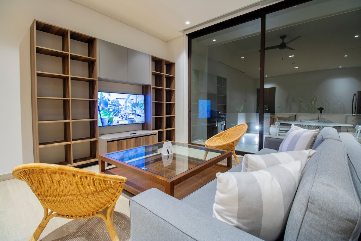 Uma sala de estar moderna com televisão de tela plana, cadeiras de vime, mesa de centro de vidro e sofá cinzento.