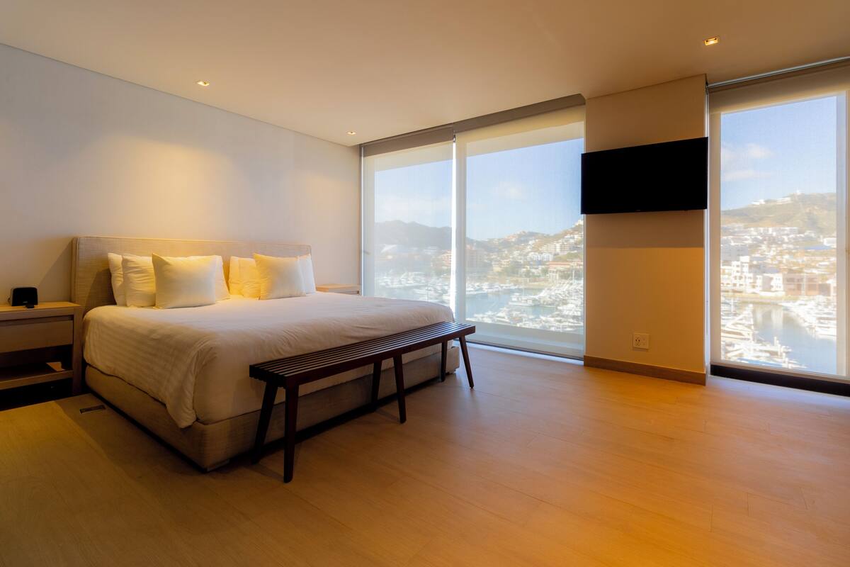 Une chambre moderne avec un grand lit, un banc, un téléviseur fixé au mur et une vue sur la ville grâce à de grandes fenêtres.