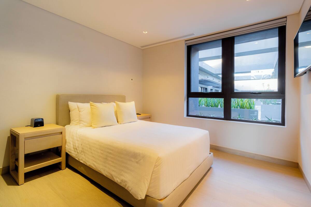Une chambre moderne avec un grand lit, une table de chevet et une fenêtre donnant sur des bâtiments et de la verdure.