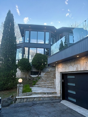 Uma casa moderna de dois andares com uma escadaria de pedra, uma garagem e uma grande árvore ao fundo.