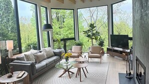 Una sala de estar moderna con sofá, sillones, mesa de centro y televisor. Amplias ventanas ofrecen vistas al verdor exterior.