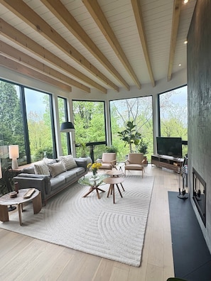 A modern living room with a sofa, chairs, a coffee table, and a TV. Large windows offer a view of greenery outside.