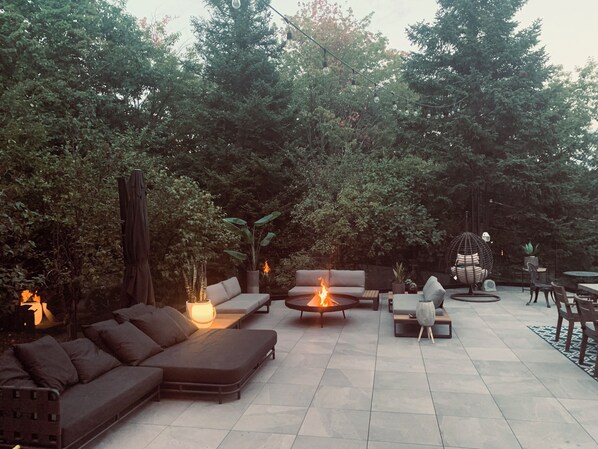 Une terrasse extérieure aménagée avec un canapé, des fauteuils, un foyer et des plantes en pot.