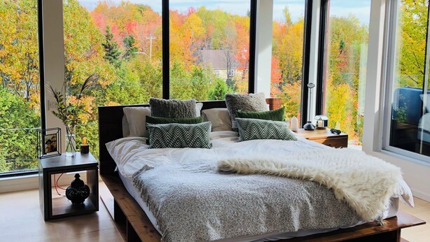 Un dormitorio moderno con una cama grande, varios ventanales y vistas al follaje otoñal.