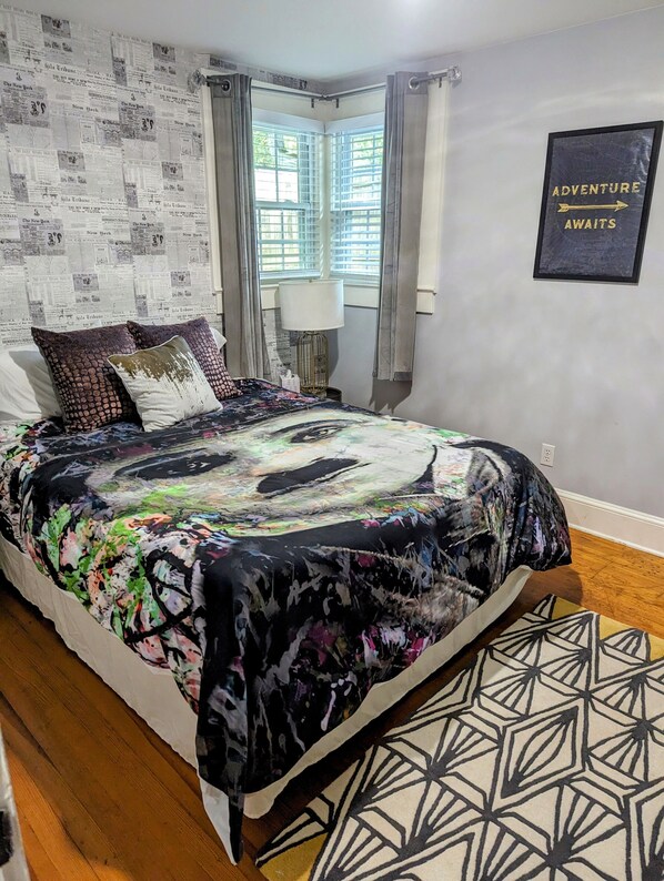 Un dormitorio con una cama cubierta con un estampado abstracto y colorido, una ventana con persianas, una lámpara y una pared con un cartel enmarcado que dice 'ADVENTURE AWAITS'.