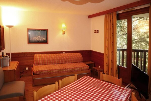 A room with a wooden bench, a table with a checkered tablecloth, a television, and a window with curtains.