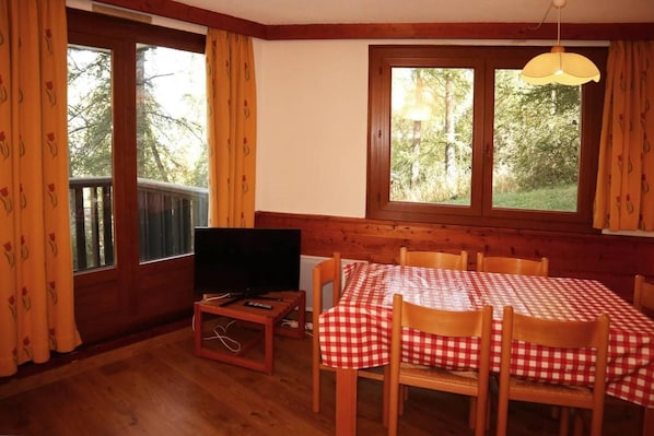 A room with a wooden table and chairs, a television on a small table, and large windows with curtains.