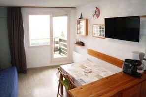 A room with a bed, a table with a coffee maker, a TV, a window with curtains, and a balcony.