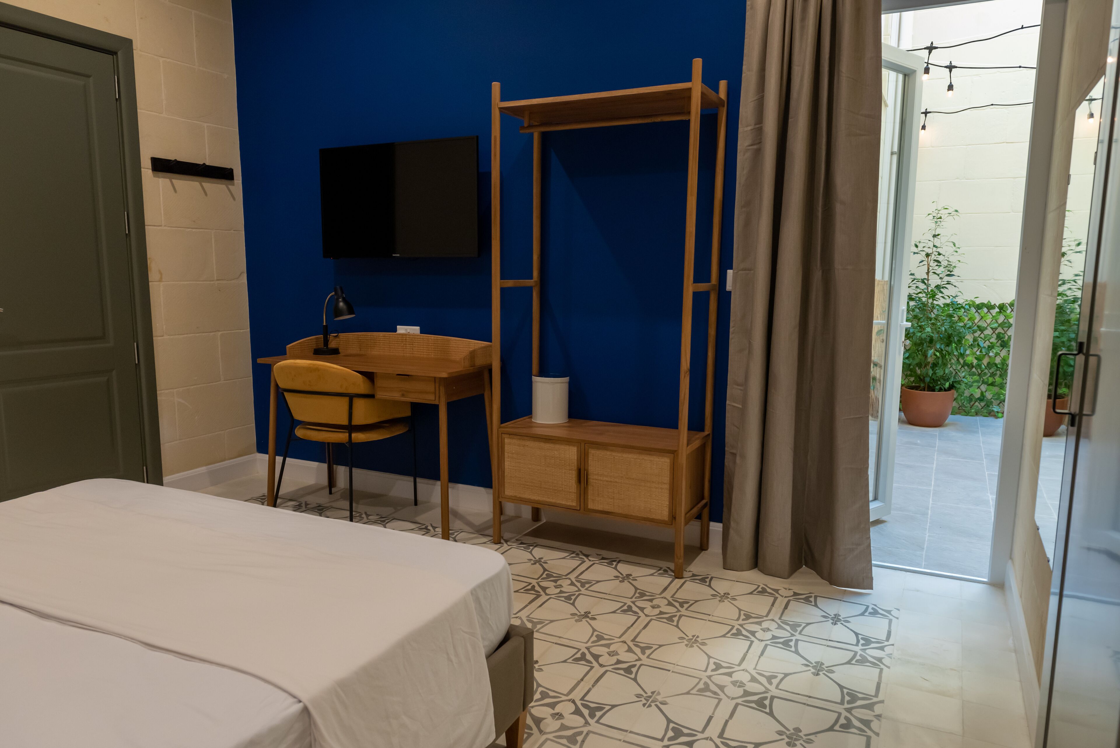 Een slaapkamer met een bed, een bureau, een stoel, een houten garderobe, een aan de muur bevestigde televisie en uitzicht buiten door een deur.