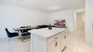 Una cucina moderna con piano di lavoro in marmo, mobili bianchi e maniglie dorate. È presente un'area pranzo con un tavolo nero e sedie blu.