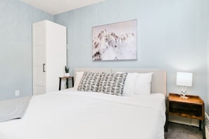 Een slaapkamer met een bed, nachtkastje met lamp, een aan de muur hangend schilderij en een deur.