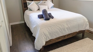 A neatly made bed with a white comforter and several pillows, a wooden headboard, and a white rug on a wooden floor.