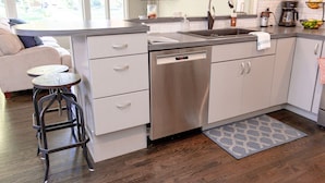A modern kitchen with a dishwasher, a coffee maker, and a fruit bowl. There are two stools and a small table.