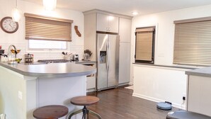 A modern kitchen with a stainless steel refrigerator, a built-in island with stools, wooden blinds, and a visible clock on the wall.