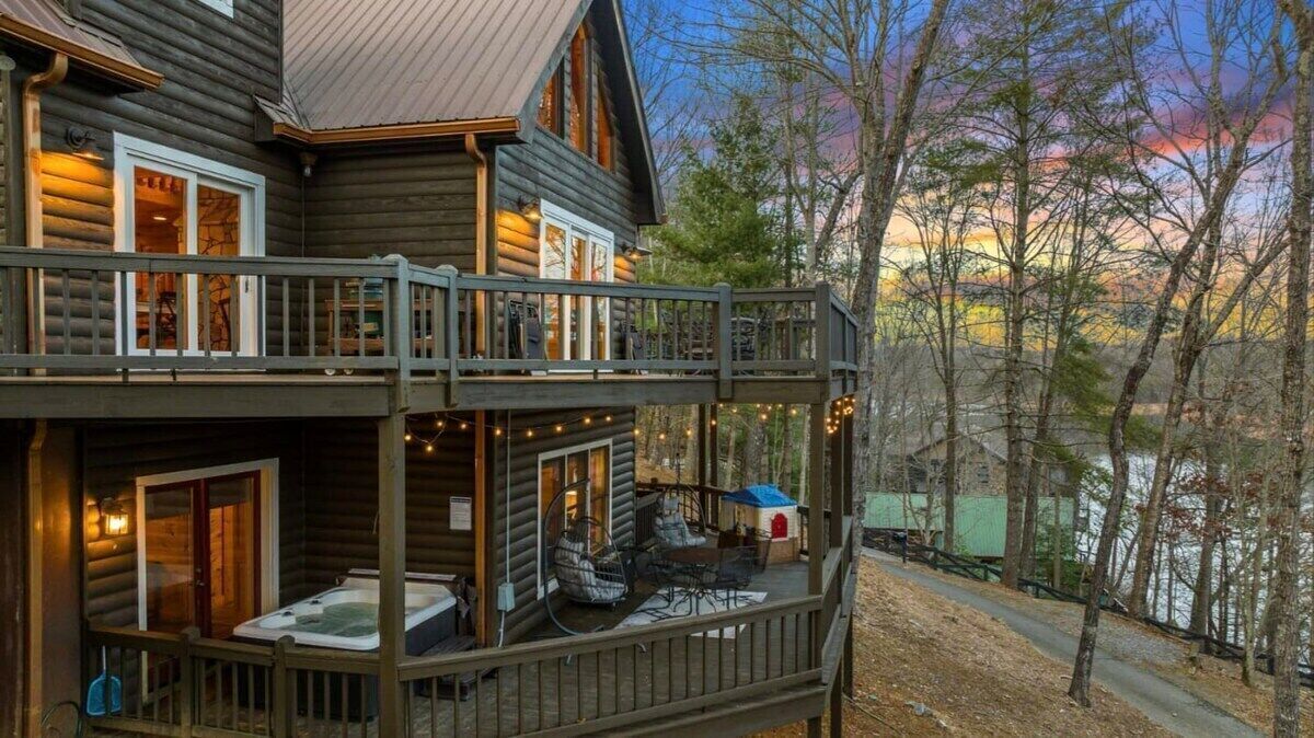 A log cabin with a deck and hot tub, surrounded by trees and a lake.