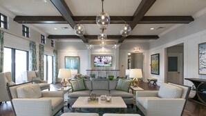 Ein großzügiges Wohnzimmer mit einer großen Eckcouch, zwei Sesseln, einem Couchtisch und einem Fernseher. Es gibt mehrere Fenster, eine Chandelierleuchtre und Kunstwerke an den Wänden.