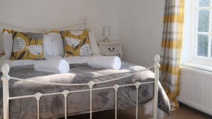A neatly made bed with a white metal frame, a bedside table with a lamp, and a window with a yellow and white checkered curtain. - Phloxhaven (Norwich)